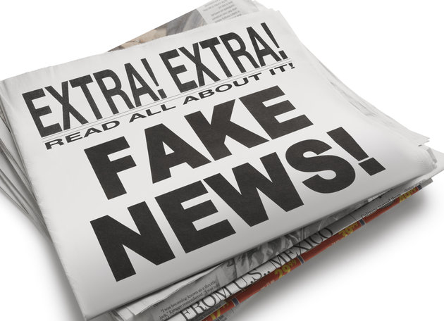 The front page of a newspaper with the headline "Fake News" which illustrates the current phenomena. Front section of newspaper is on top of loosely stacked remainder of newspaper. All visible text is authored by the photographer. Photographed in a studio setting on a white background with a slight wide angle lens.