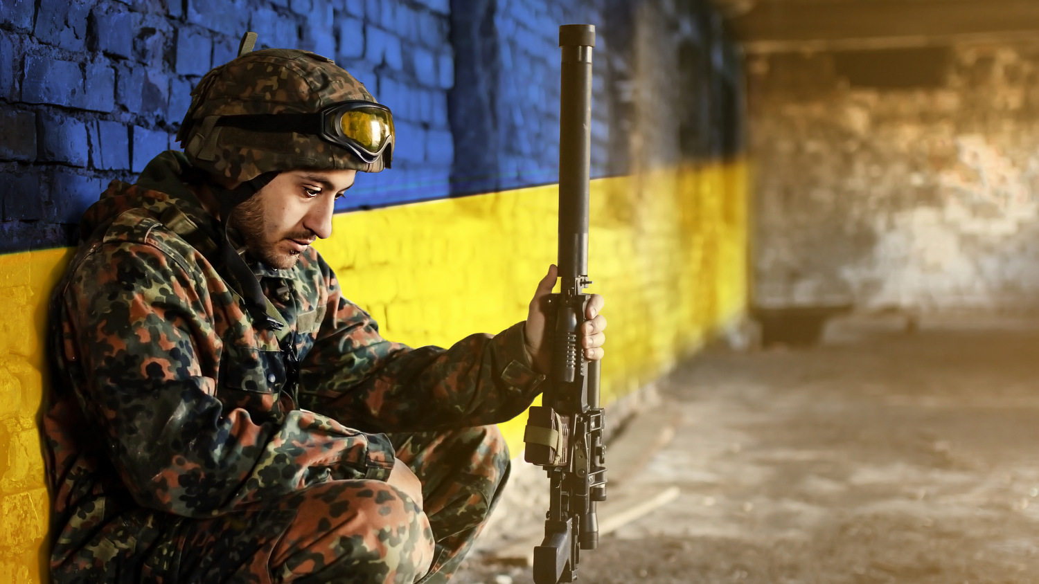 Military soldier on the background of the flag of Ukraine. The flag of Ukraine is painted on a brick wall, a tired sad soldier sits with a weapon in his hands. Relations between Ukraine and Russia